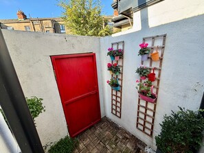 Une ruelle étroite avec une porte rouge, des plantes en pot et un treillage en bois adossé à un mur blanc.