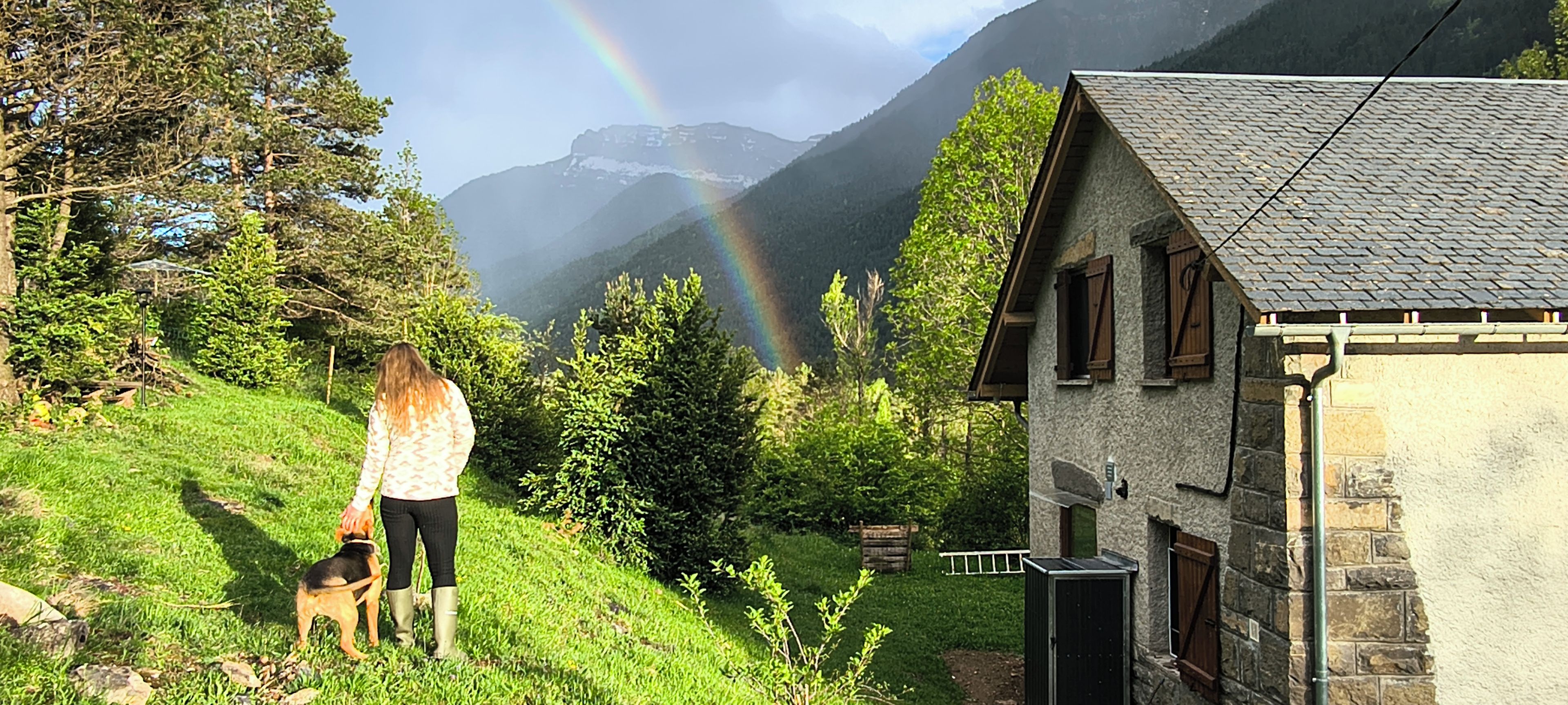 Een persoon met een hond op een grashelling, een stenen gebouw en een berg waarop een regenboog te zien is.