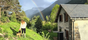 A person with a dog on a grassy slope, a stone building, and a mountain with a rainbow in the background.