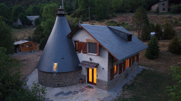 A stone house with a conical roof and a rectangular extension, surrounded by a rural landscape.