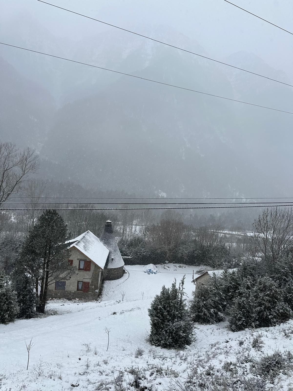 Paesaggio innevato con una casa e una struttura conica, forse un silo, circondato da alberi e una montagna sullo sfondo.