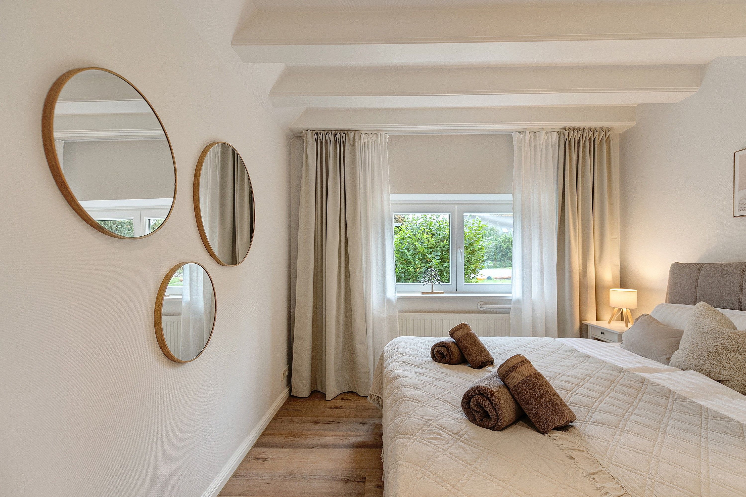 Ein Schlafzimmer mit einem Bett, zwei Spiegeln, einem Fenster mit Vorhängen und einem Nachttisch mit einer Lampe.