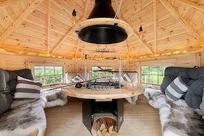 A cozy cabin interior with a central wood-burning stove, a dining table, and two sofas.