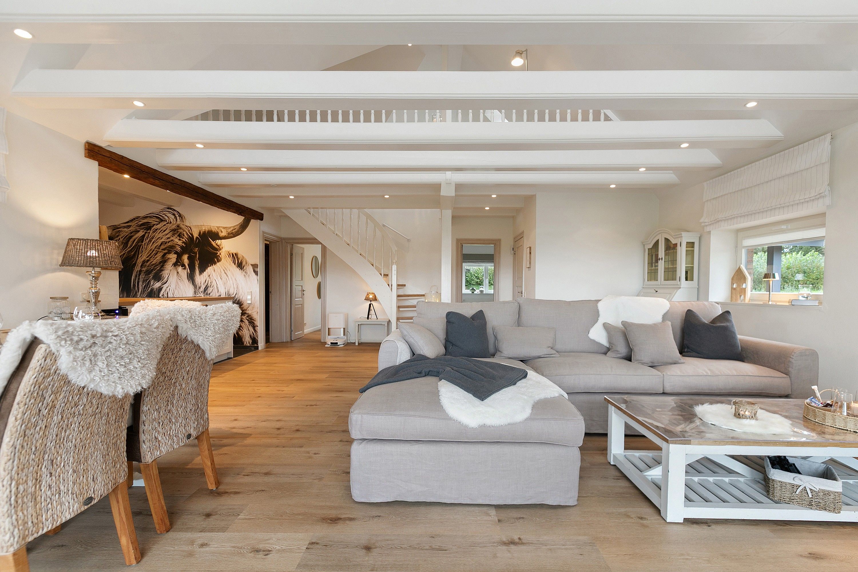 Un salón espacioso con un amplio sofá seccional gris, una mesita de centro y una silla de fibra natural. Una escalera con barandilla de madera conduce a un nivel superior.