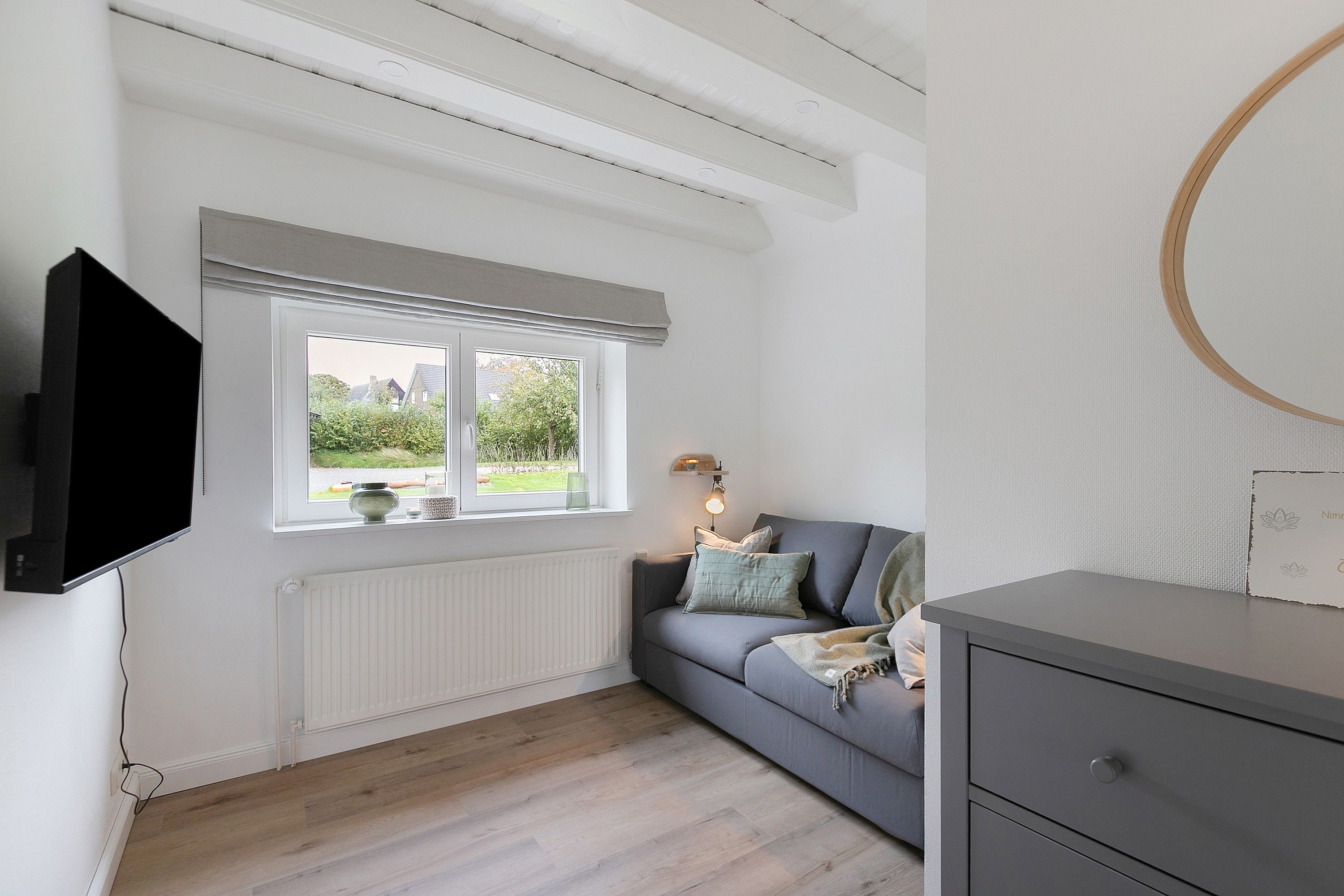 Une pièce avec un canapé gris, un téléviseur fixé au mur, une fenêtre donnant sur un paysage verdoyant et un radiateur placé sous la fenêtre.