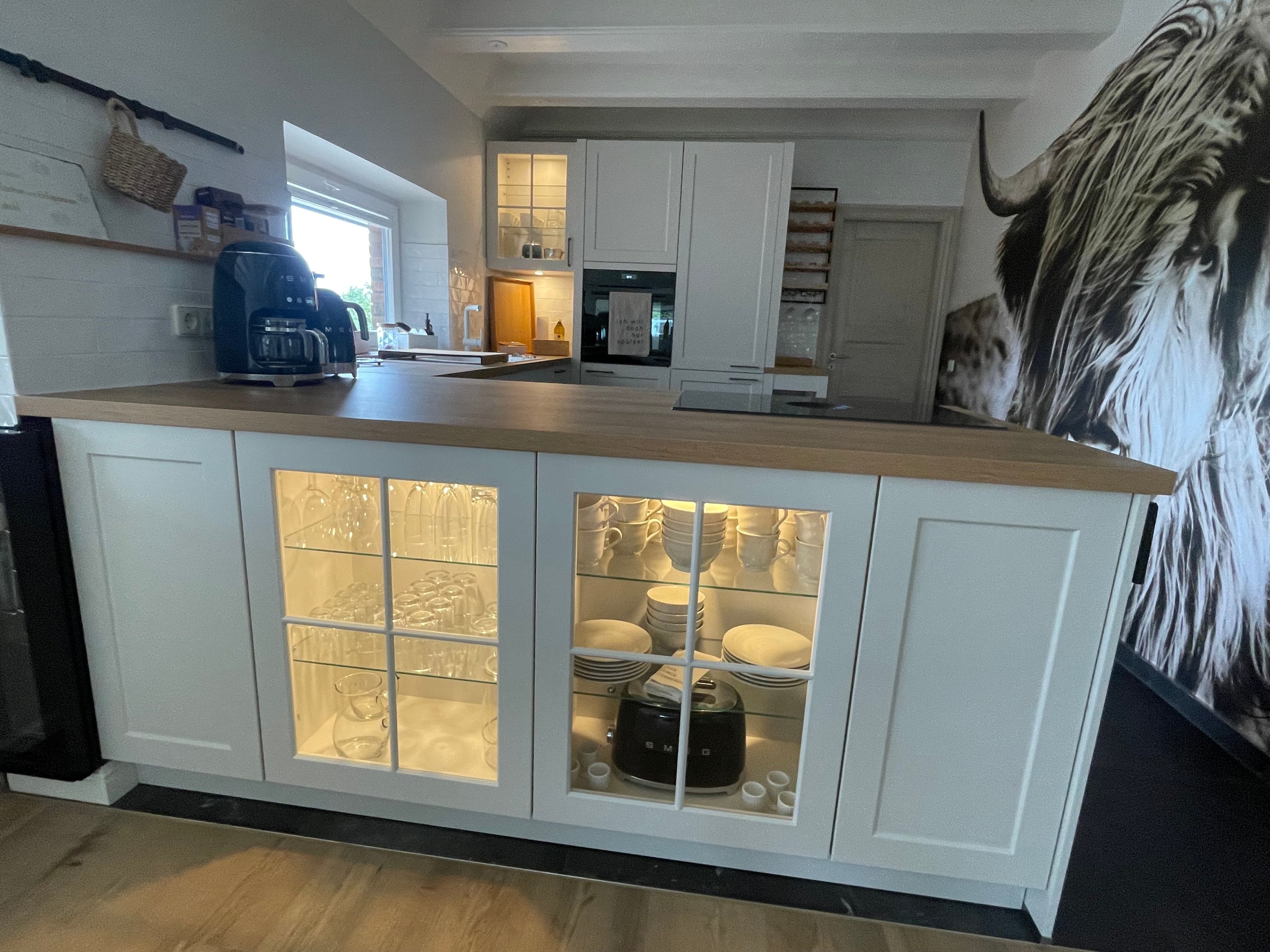 Une cuisine moderne avec des meubles blancs, un plan de travail en bois et des placards à façade en verre pour ranger la vaisselle et les ustensiles.