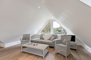 A modern loft bedroom with a sloped ceiling, a large window, and a cozy seating area with a sofa and chairs.