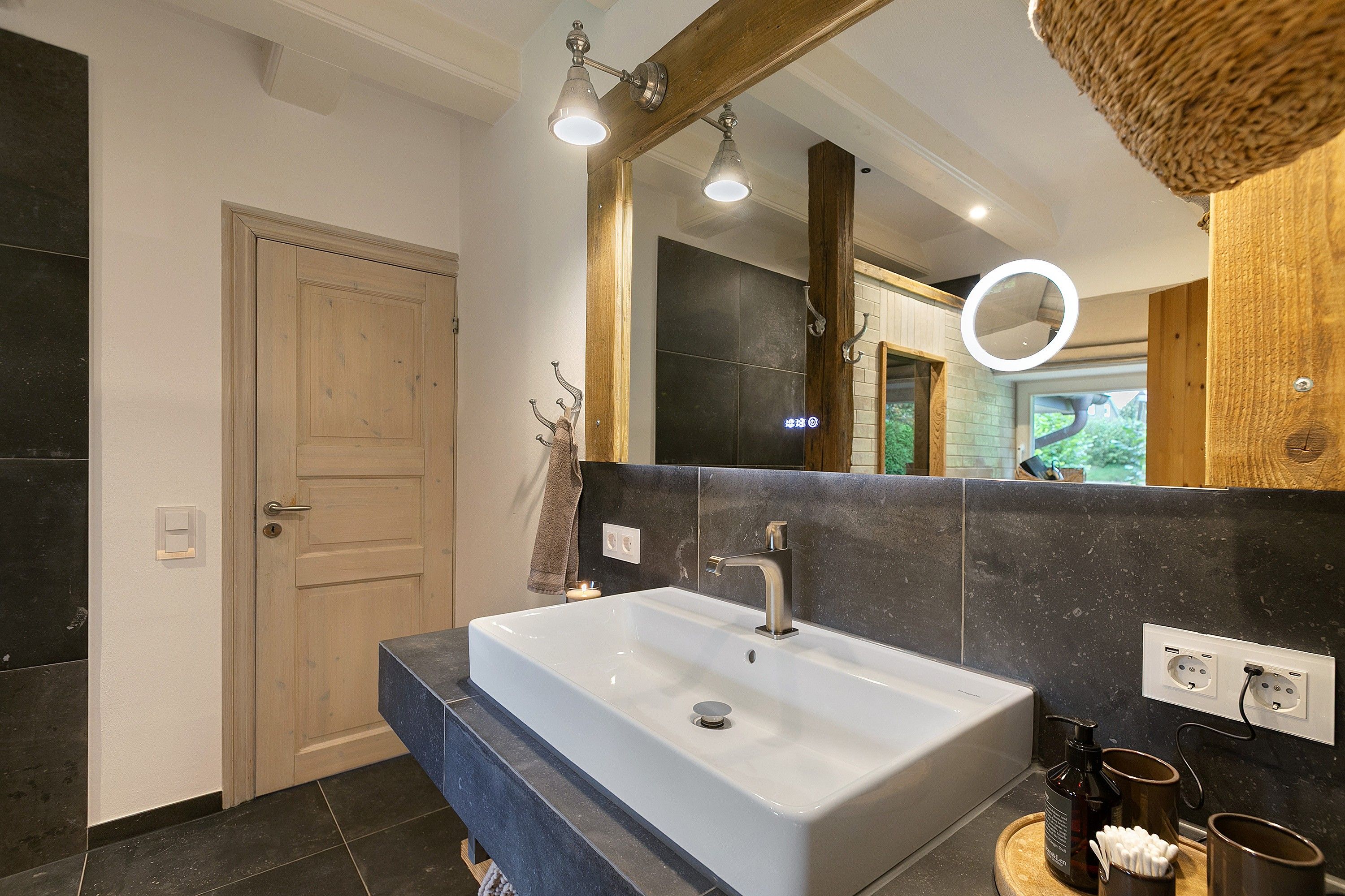 A modern bathroom with a large mirror, a white sink, and a wooden door.