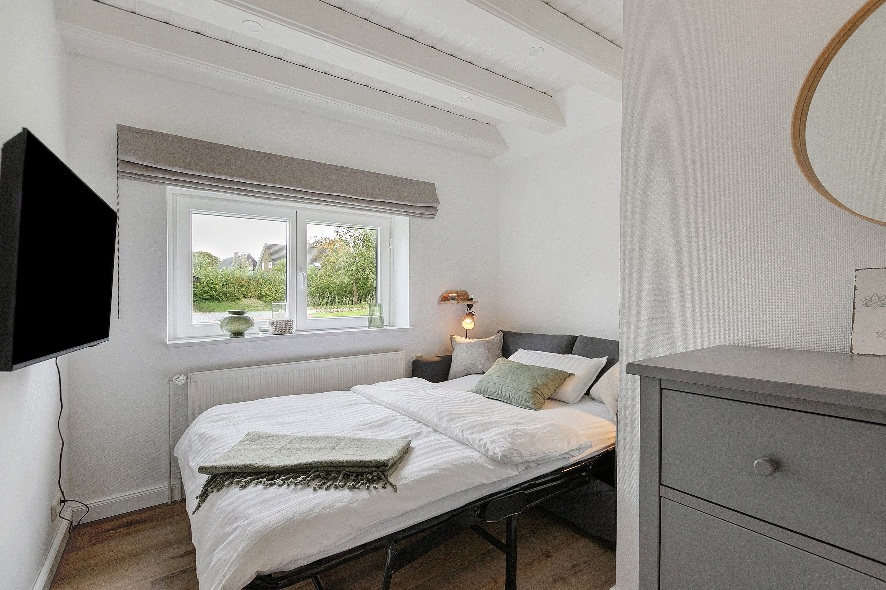A bedroom with a bed, bedside table, lamp, and a window with a view of greenery.