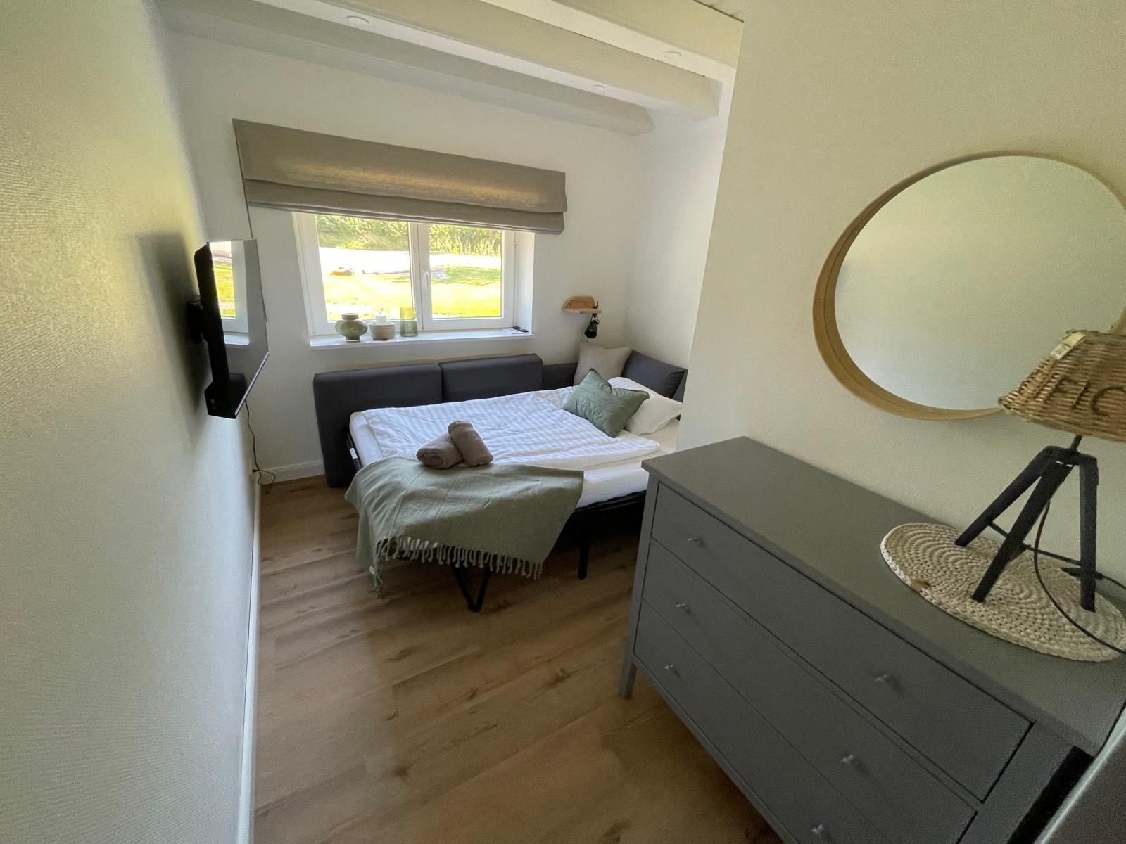 Een slaapkamer met een bed, nachtkastje, lamp en een raam met uitzicht op groen.