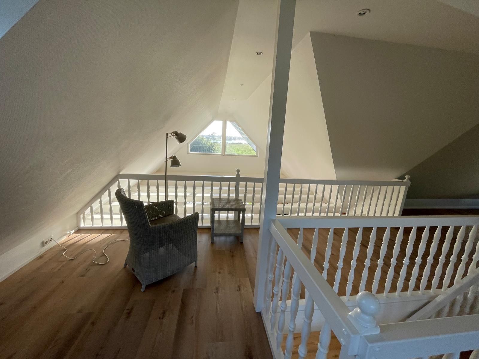 A spacious attic room with a wooden floor, a white railing, and a large window providing a view of the outdoors.