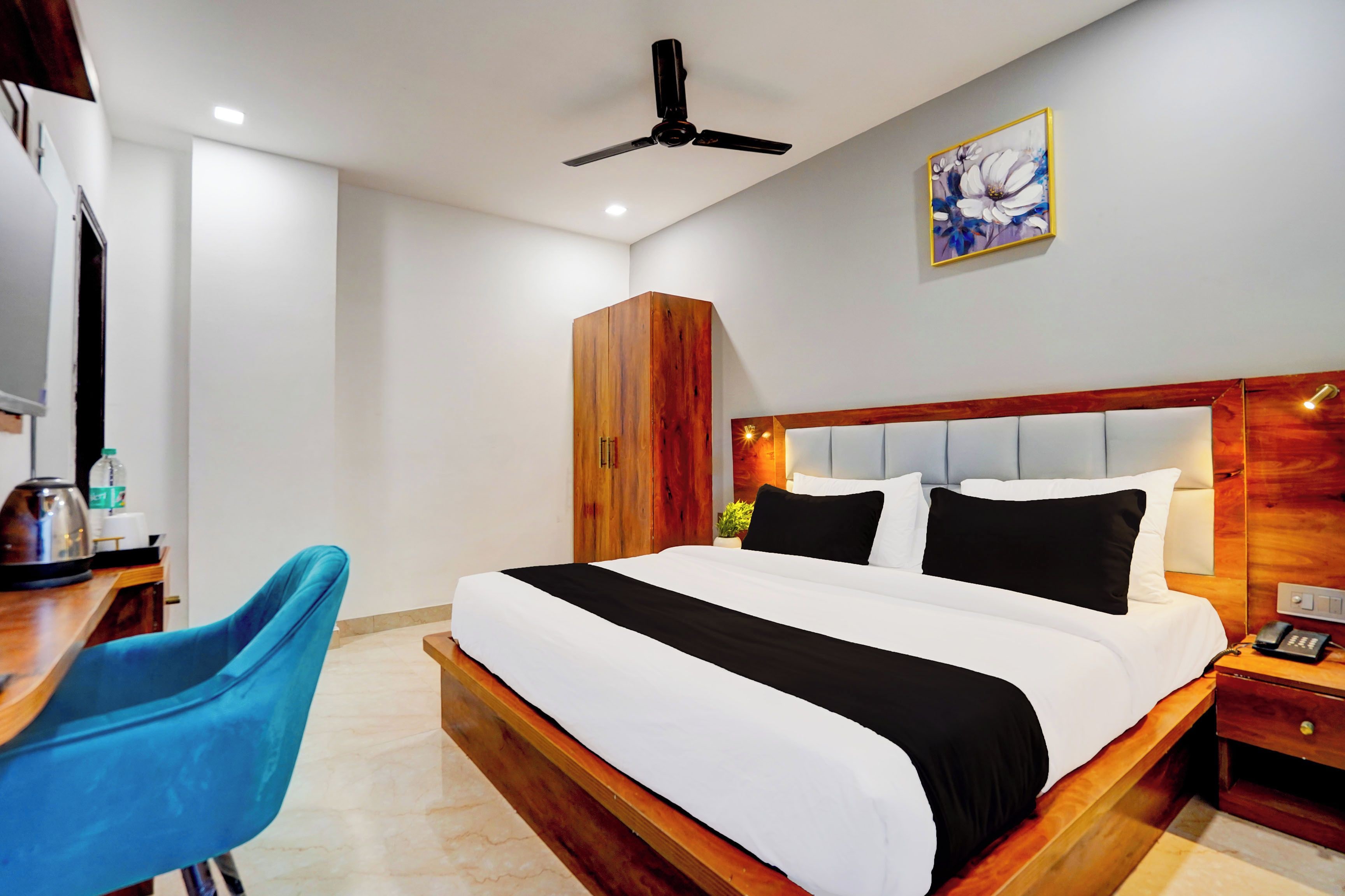A hotel room with a bed, a desk, a chair, a wardrobe, a ceiling fan, and a painting on the wall.
