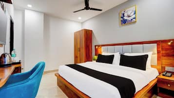 A hotel room with a bed, a desk, a chair, a wardrobe, a ceiling fan, and a painting on the wall.