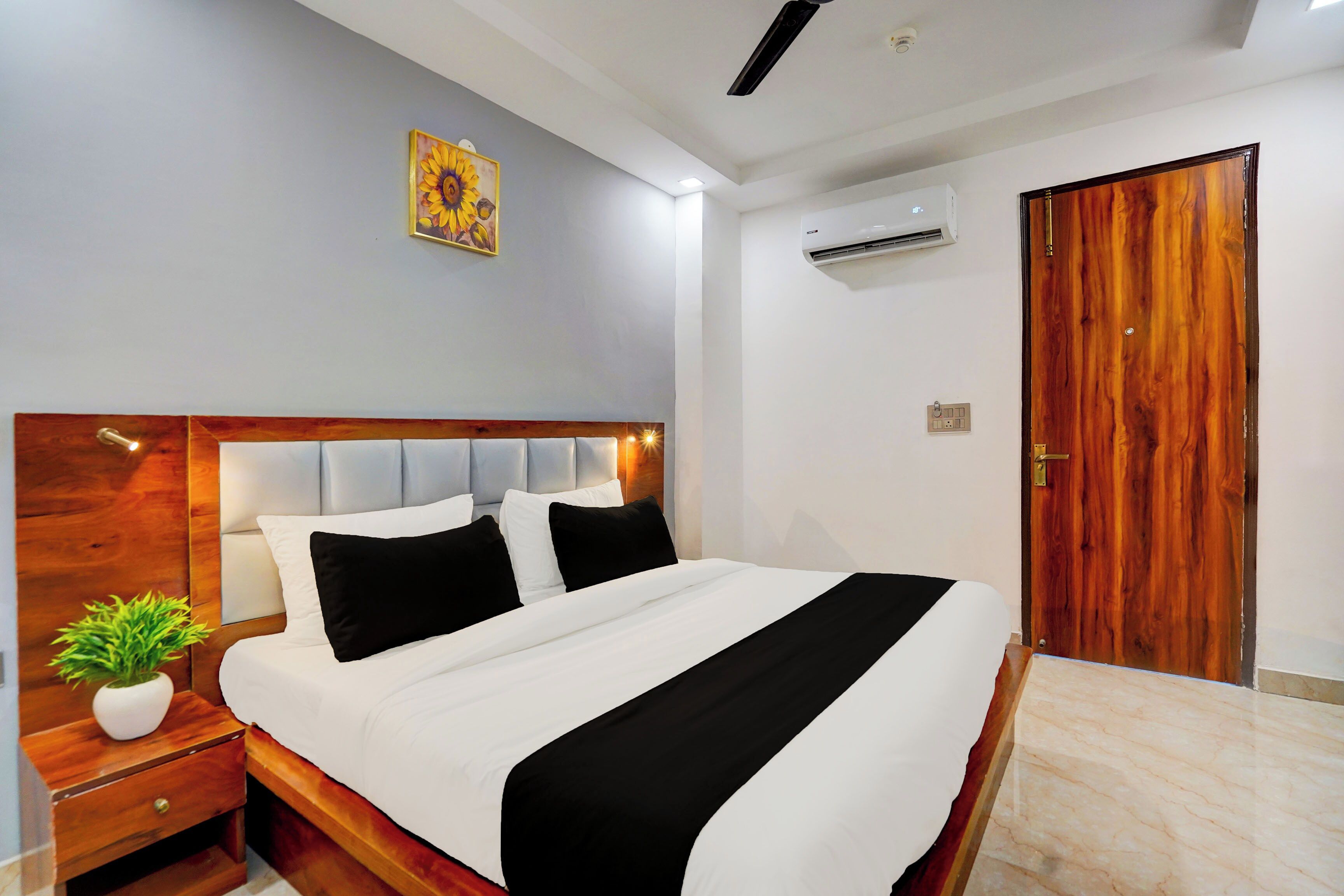 A hotel room with a wooden headboard, a bed with white and black bedding, a nightstand with a plant, and a wooden door.
