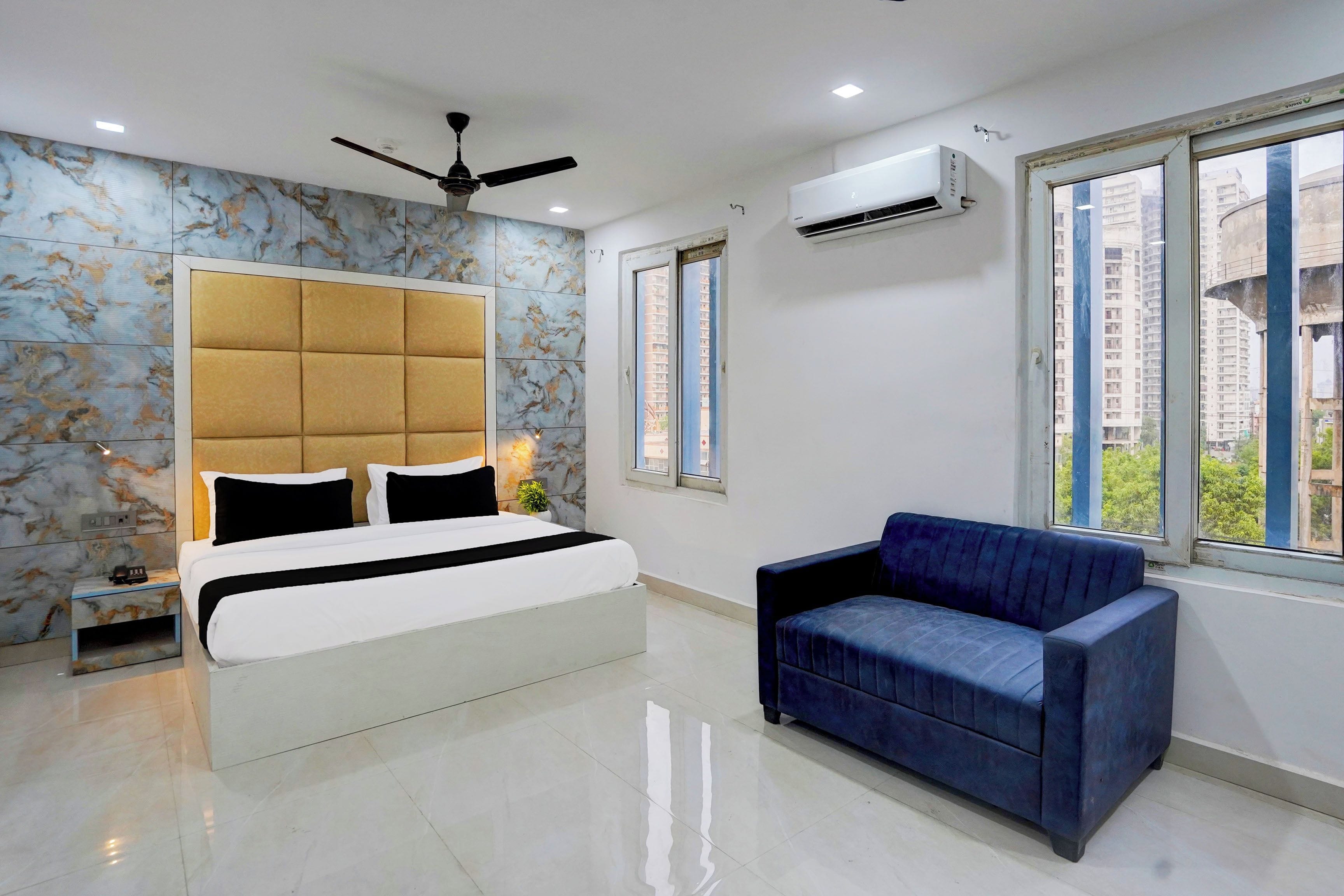 Un dormitorio con una cama, un sofá azul, un ventilador de techo, un equipo de aire acondicionado y una ventana con vistas a edificios.