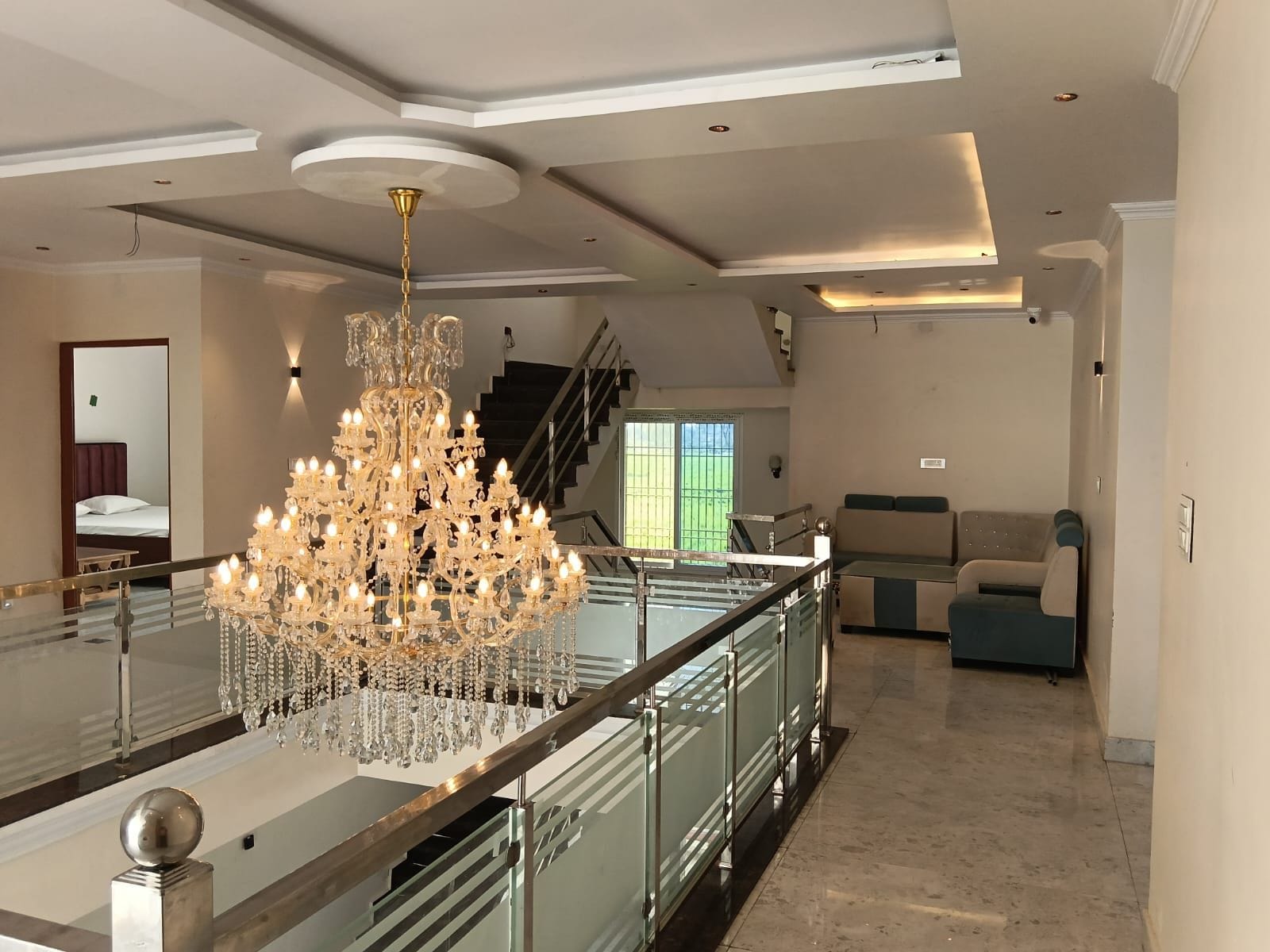 A spacious living area with a grand chandelier, a staircase, and a view of a green outdoor area.