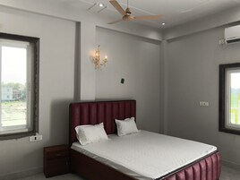 Un dormitorio con cama, una mesita de noche, aire acondicionado instalado en la pared, ventilador de techo y ventana con vistas a edificios.