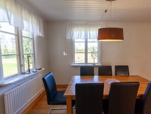 A dining area with a wooden table and chairs, large windows with white curtains, and a hanging light fixture. - Holiday Home With 4 Bedrooms (Ambjörnarp)