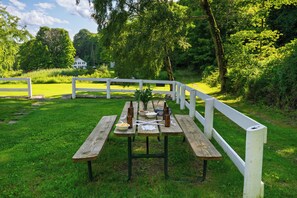 Ein Picknicktisch mit Speisen und Getränken, umgeben von einem weißen Zaun in einer grünen Umgebung mit Bäumen.