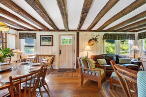 Ein rustikaler Innenraum mit Holzbalken, einem Essbereich mit Stühlen und Tisch, einer Couch mit Kissen und einem Hirschkopf an der Wand.