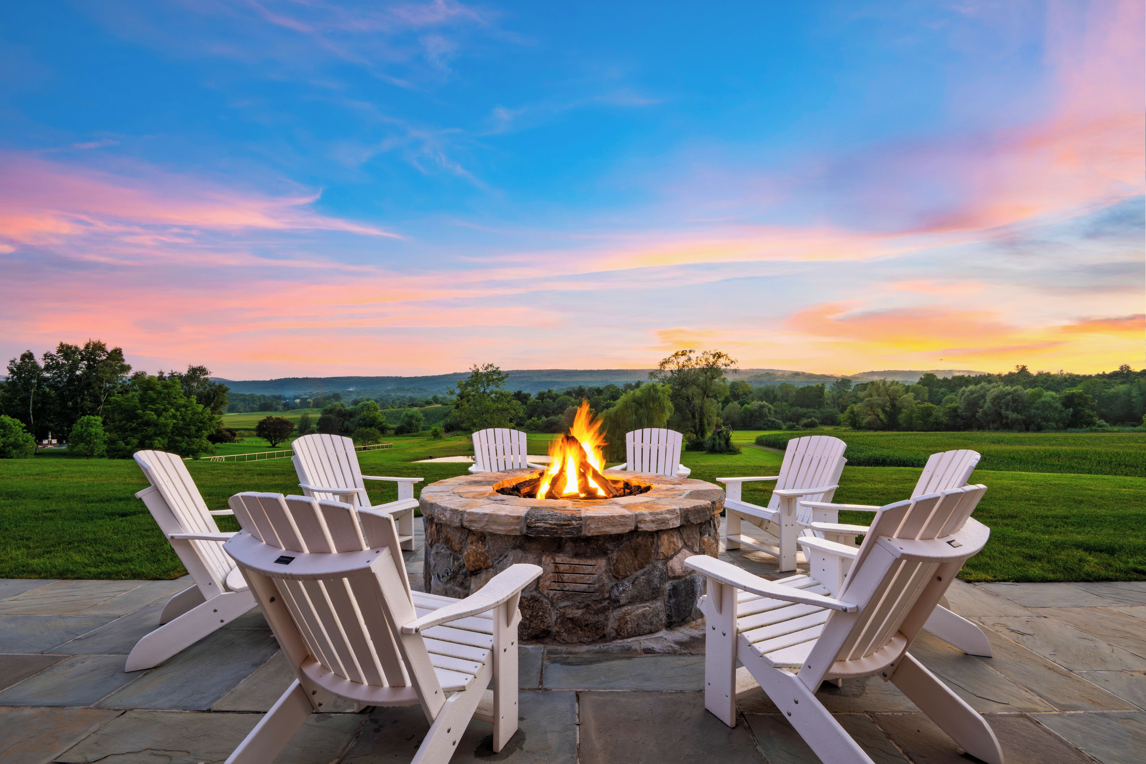Ein Steinkamin mit Feuer, umgeben von weißen Adirondack-Stühlen, auf einer Terrasse mit Blick auf eine grüne Landschaft und Hügel.