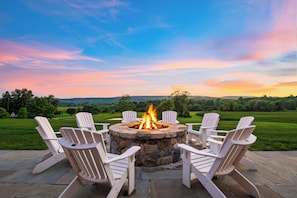 Ein Steinkamin mit Feuer, umgeben von weißen Adirondack-Stühlen, auf einer Terrasse mit Blick auf eine grüne Landschaft und Hügel.
