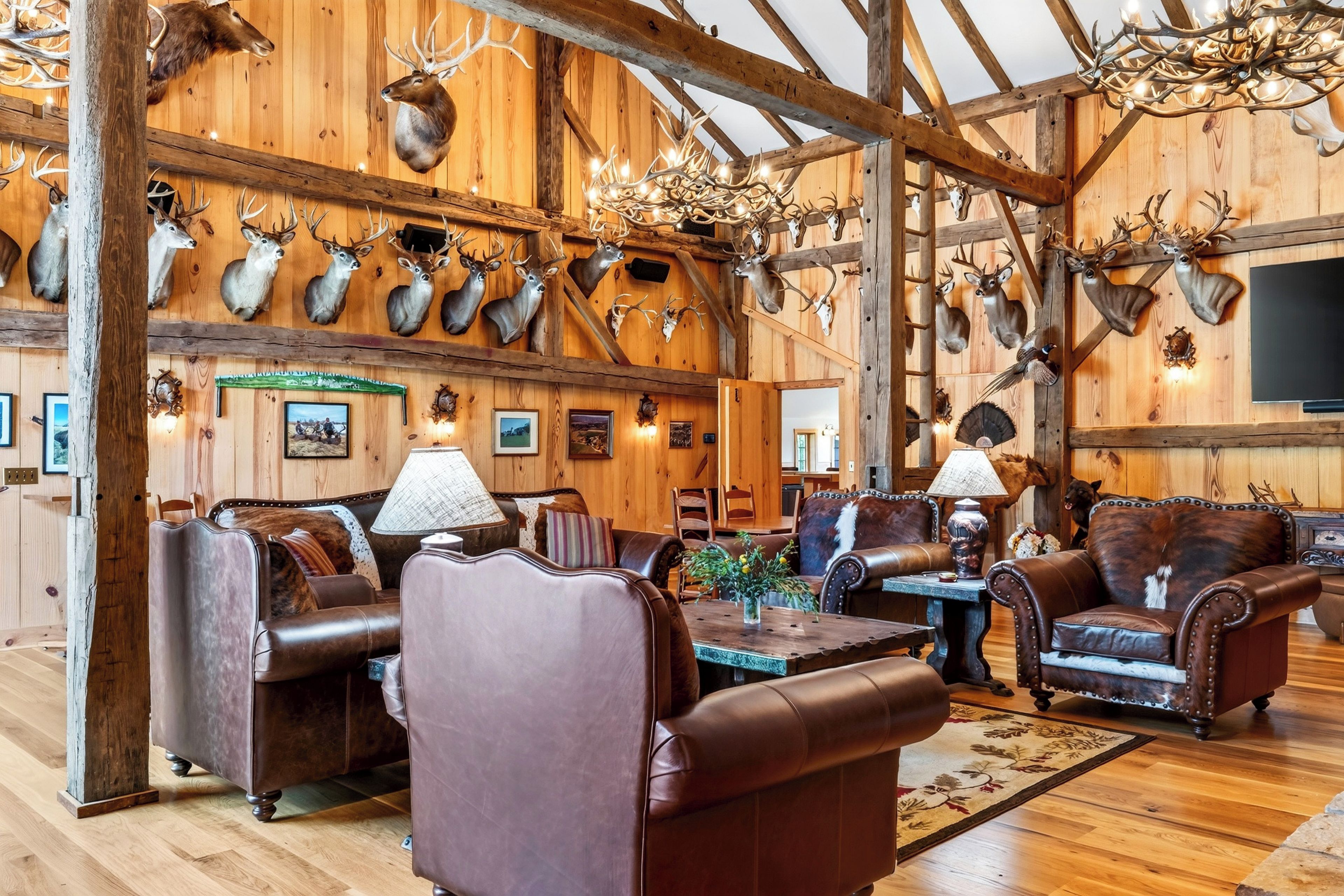 A rustic interior with wooden walls, mounted animal heads, leather furniture, and a chandelier.