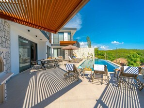 Ein moderner Poolbereich im Freien mit Liegestühlen und einem Esstisch, umgeben von einer Steinmauer und einer Holzpergola.
