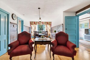 A room with two red patterned chairs, a wooden table, and a dining area with a chandelier. - Entire 1750 Inn + Event Barn + Concierge | Old Drovers Inn & Carriage House (Dover Plains)