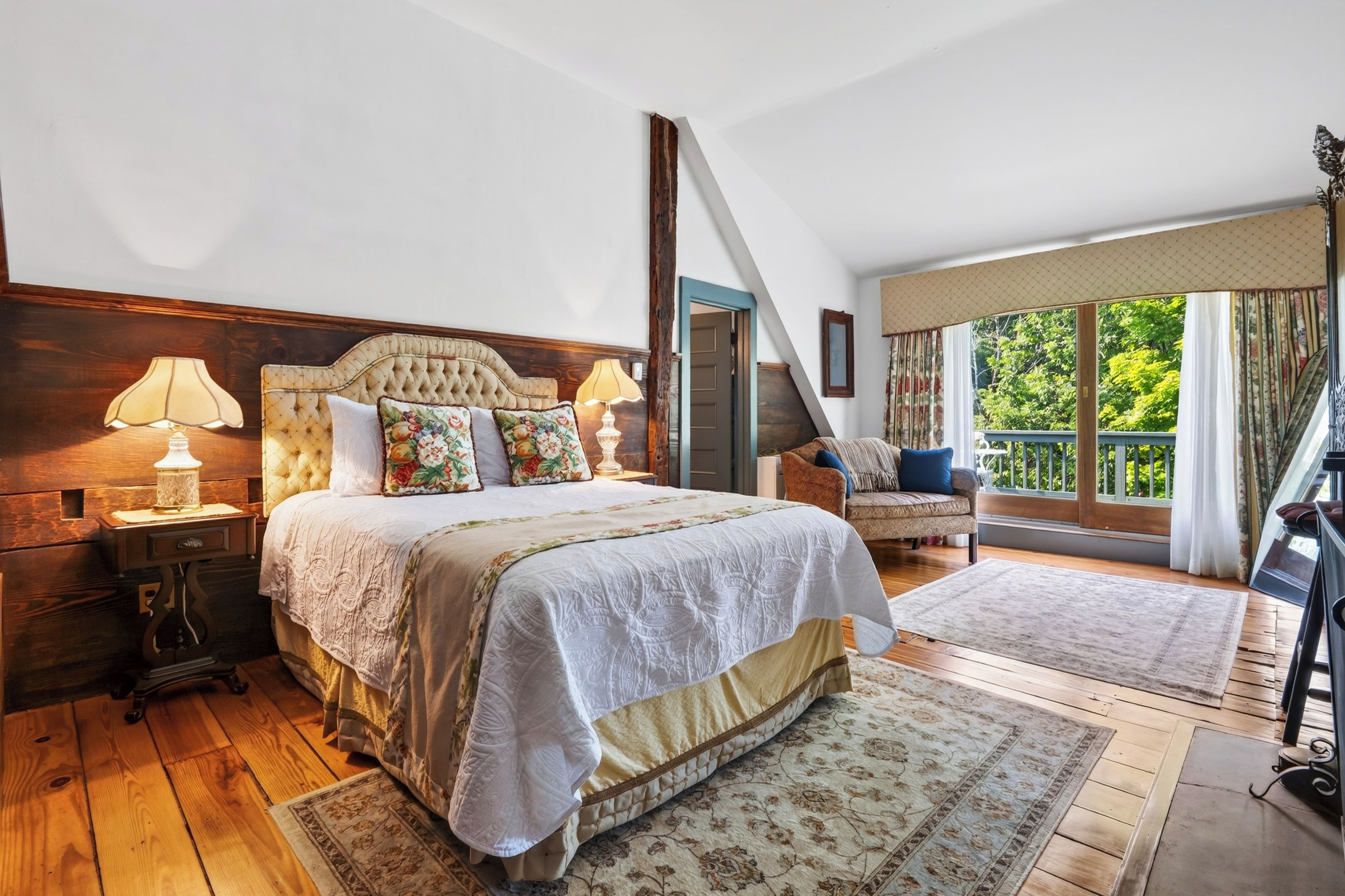 A bedroom with a large bed, a nightstand, a lamp, a chair, and a view of greenery through the window.
