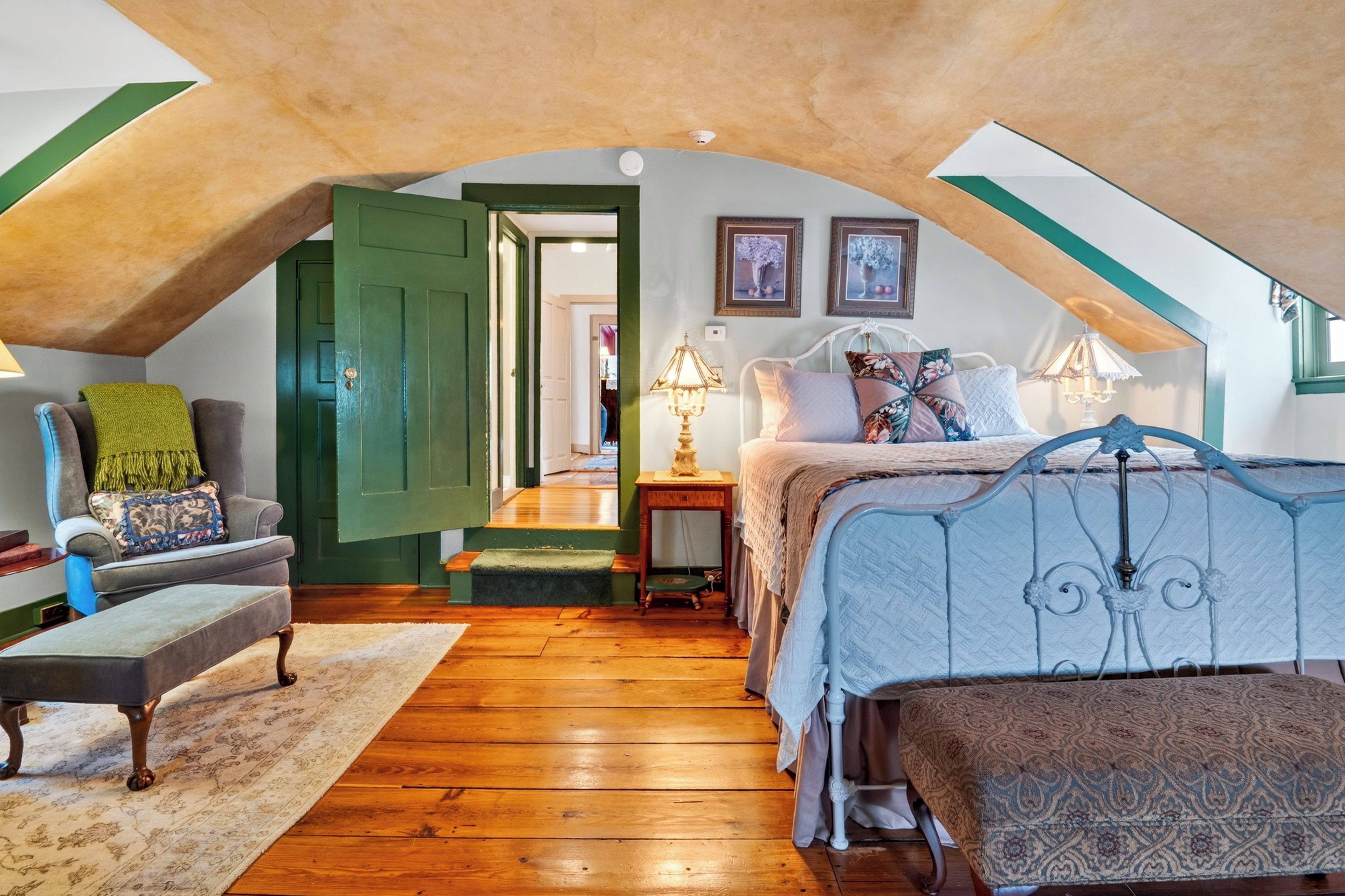 Une chambre mansardée chaleureuse, avec un plafond en pente, un lit doté d’une tête de lit à motifs, un banc, une chaise et une petite table avec une lampe.