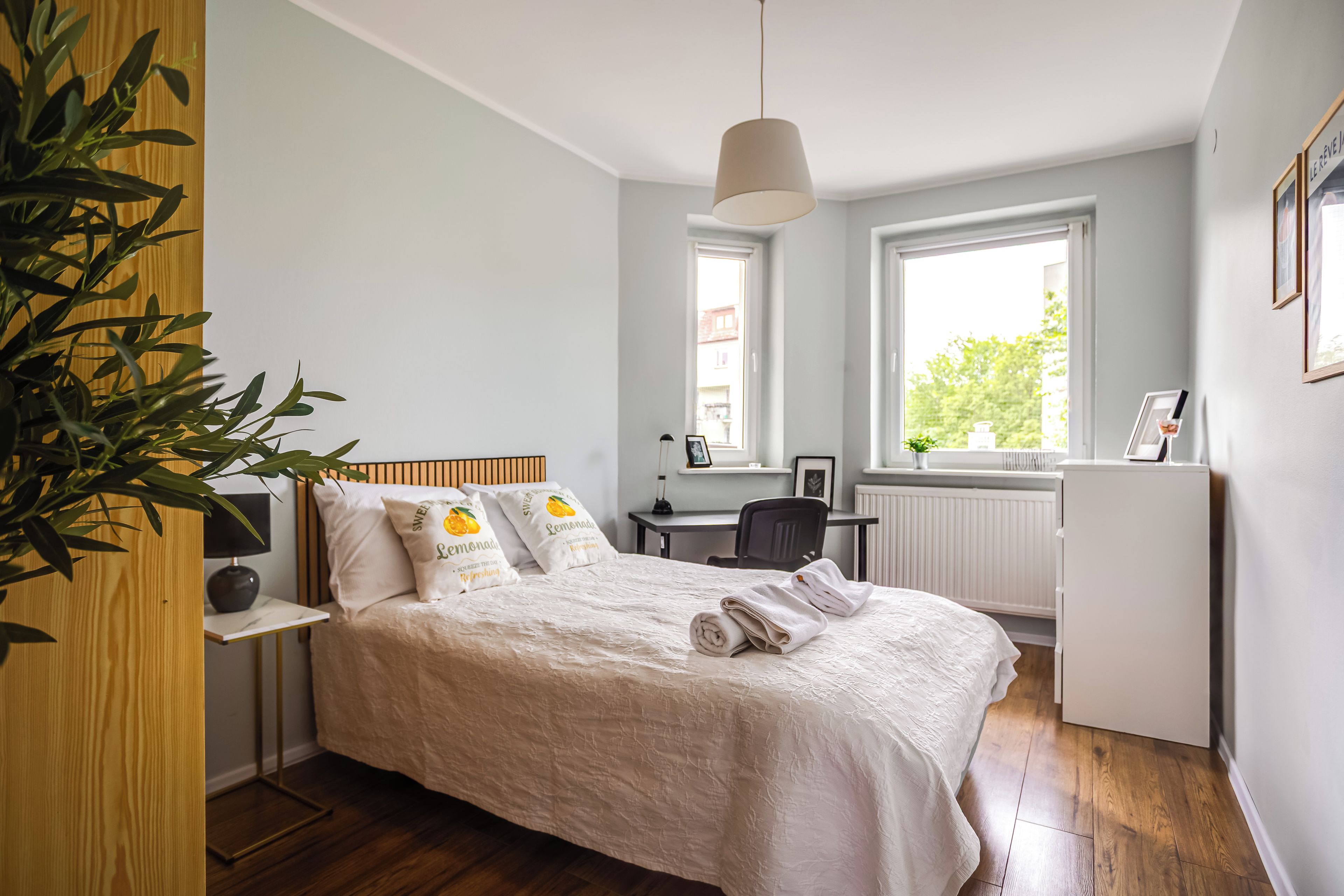 Un dormitorio con cama, mesita de noche, lámpara, escritorio, silla, radiador y ventanas.