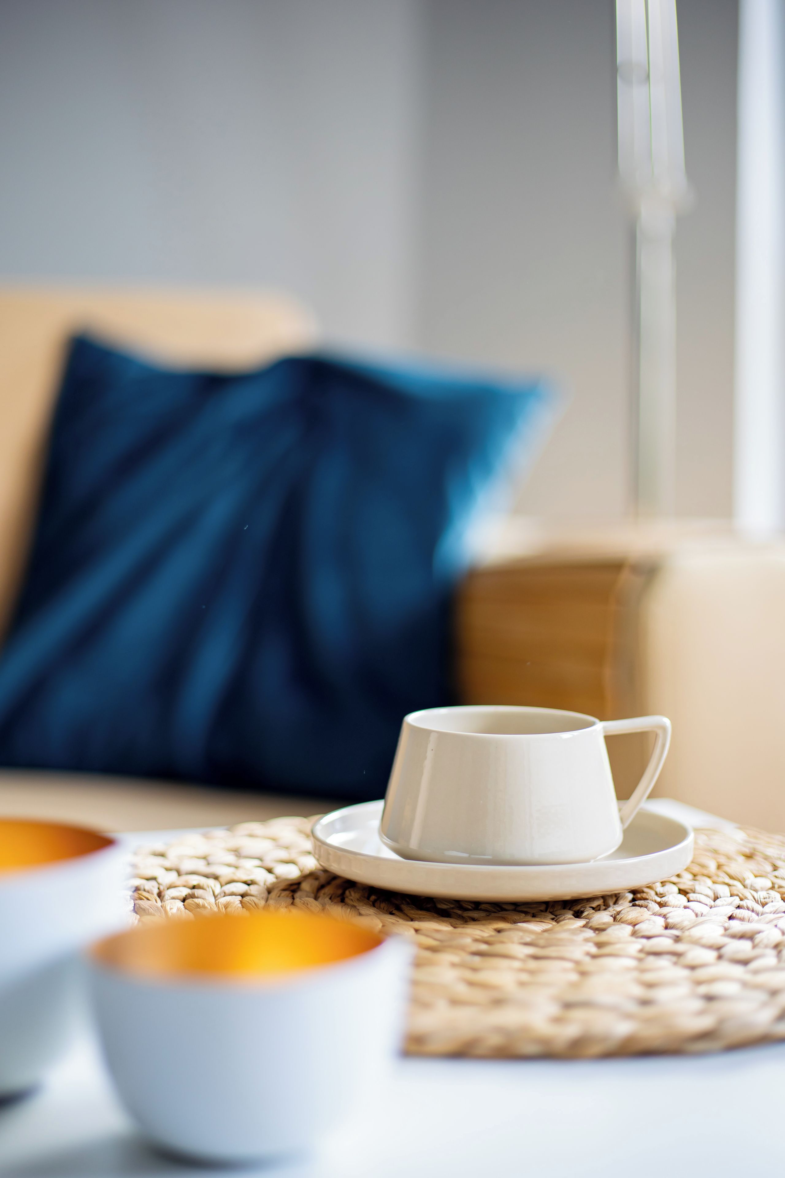 Una taza blanca sobre un platito, sobre una base tejida, con un tazón de cereal al frente y una almohada azul al fondo.