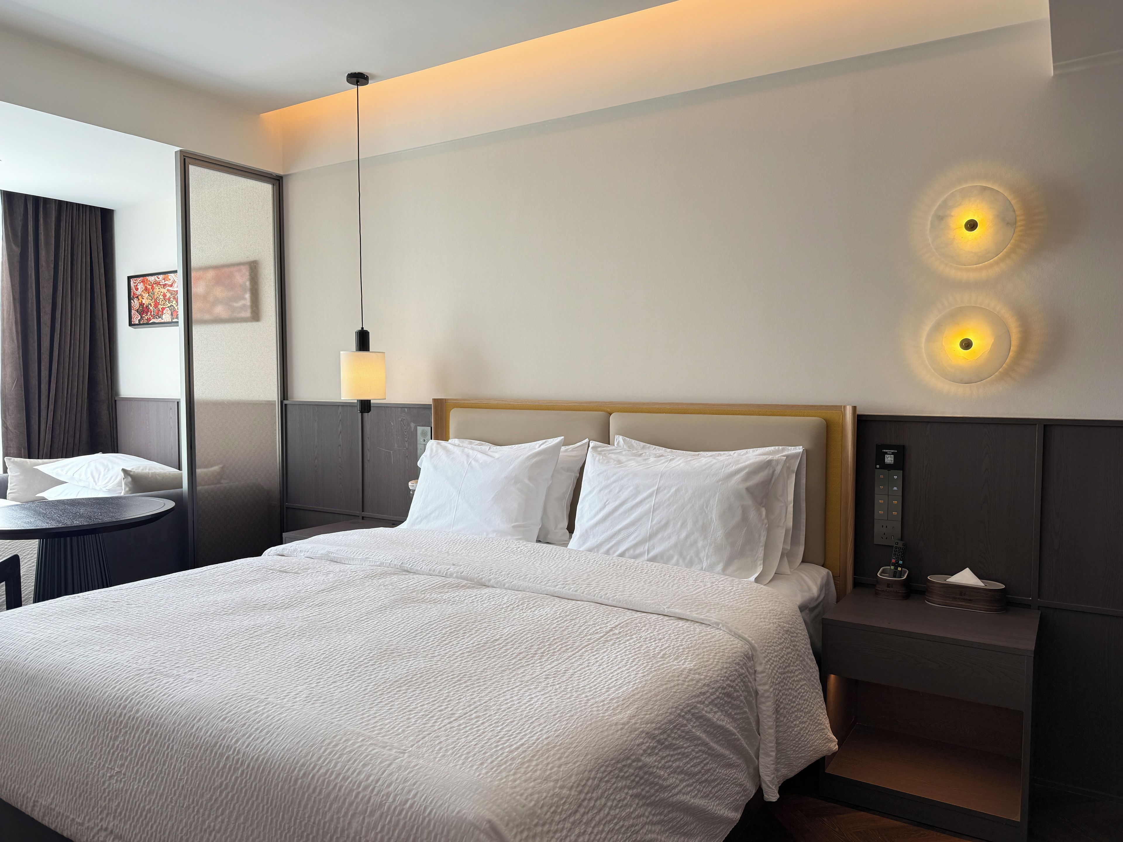 Et hotelværelse med seng, sengebord, lampe og vægmonteret lys.