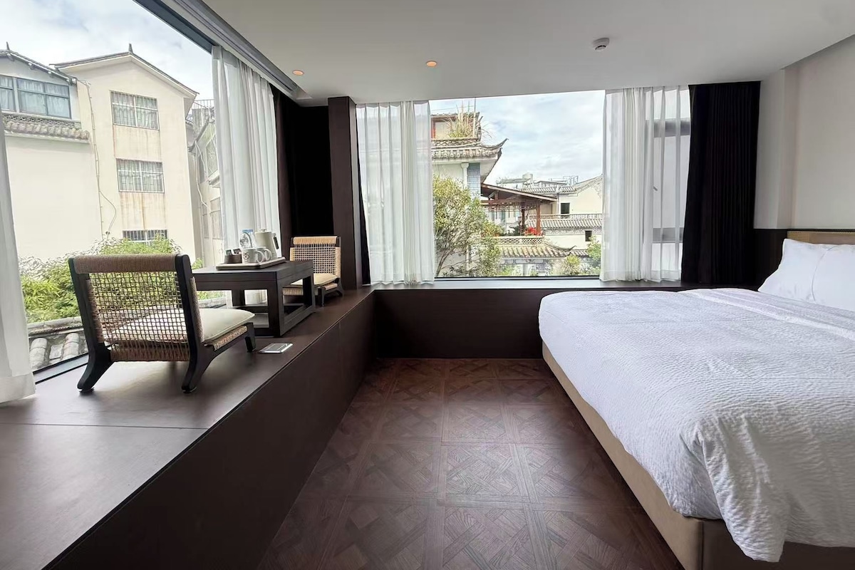 A hotel room with a bed, a small table with chairs, and a view of buildings through large windows.