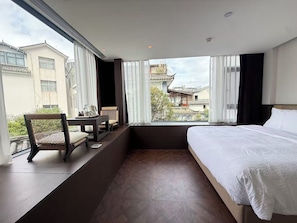 A hotel room with a bed, a small table with chairs, and a view of buildings through large windows. - Wanda Moments Resort Dali Ancient City (Dali)