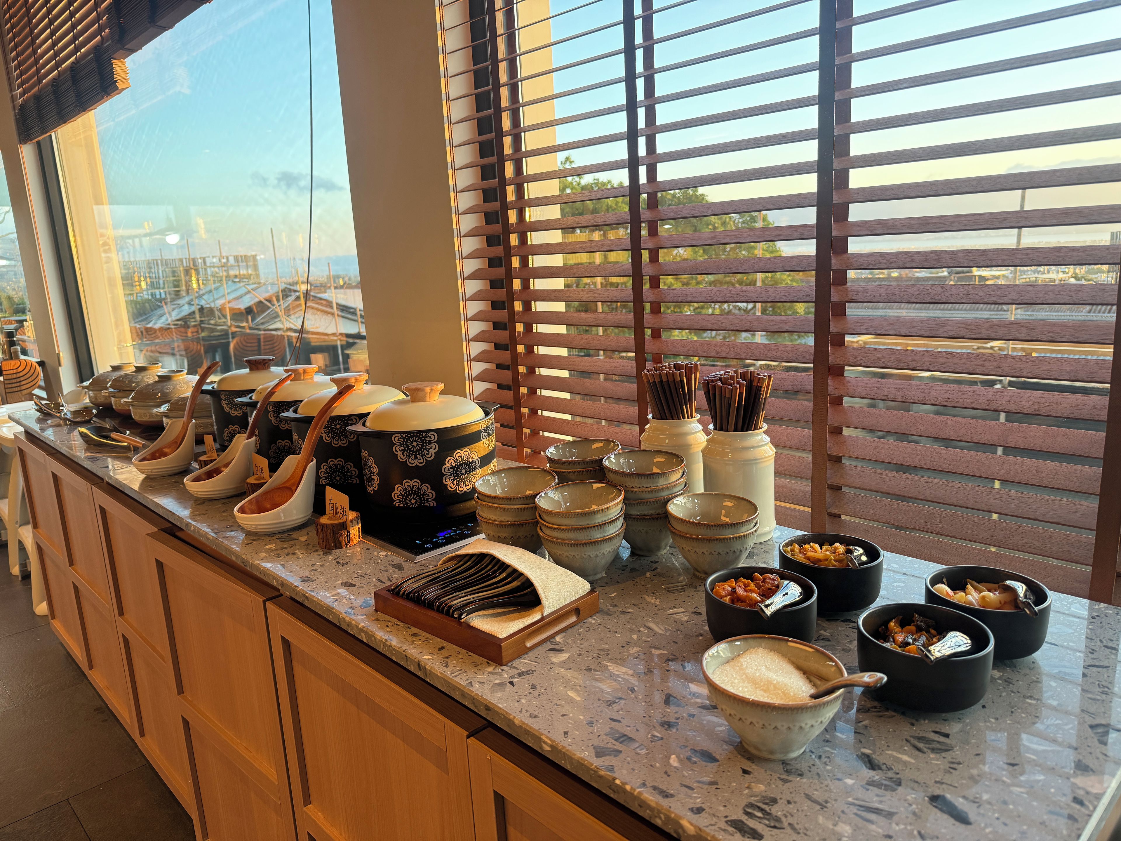 Una encimera de cocina con varios boles y utensilios, una ventana con persianas, y vista a un edificio.
