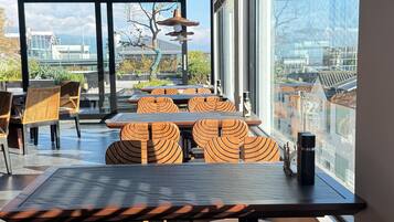 Un espace de repas moderne, doté d’une grande table et de chaises, de larges fenêtres offrant une vue sur les bâtiments et les arbres, et d’un plafond orné d’un motif tissé.