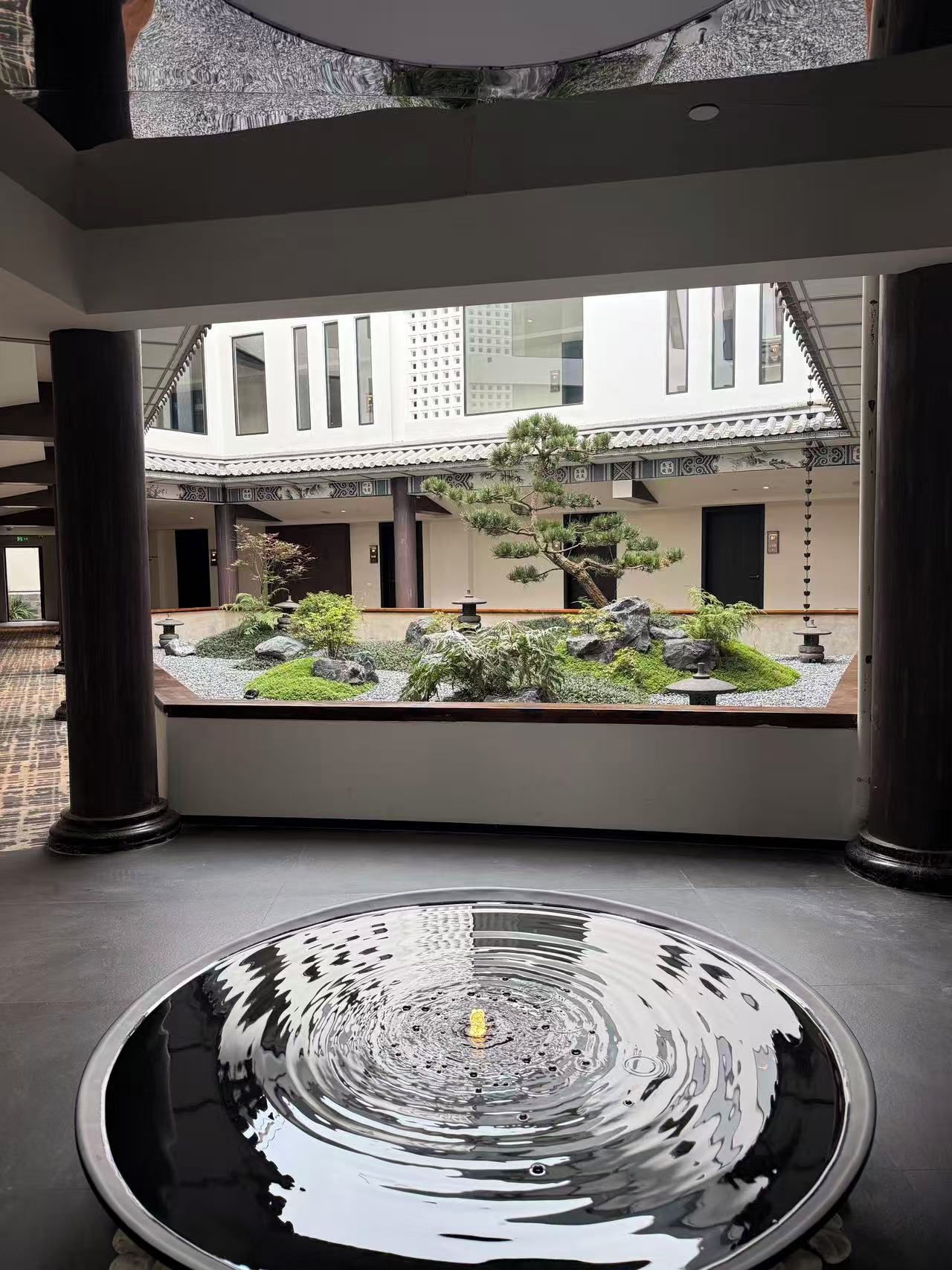 Un jardin intérieur paisible, agrémenté d’un élément aquatique, présentant une architecture de style japonais traditionnel et offrant une vue sur un bâtiment aux grandes fenêtres.