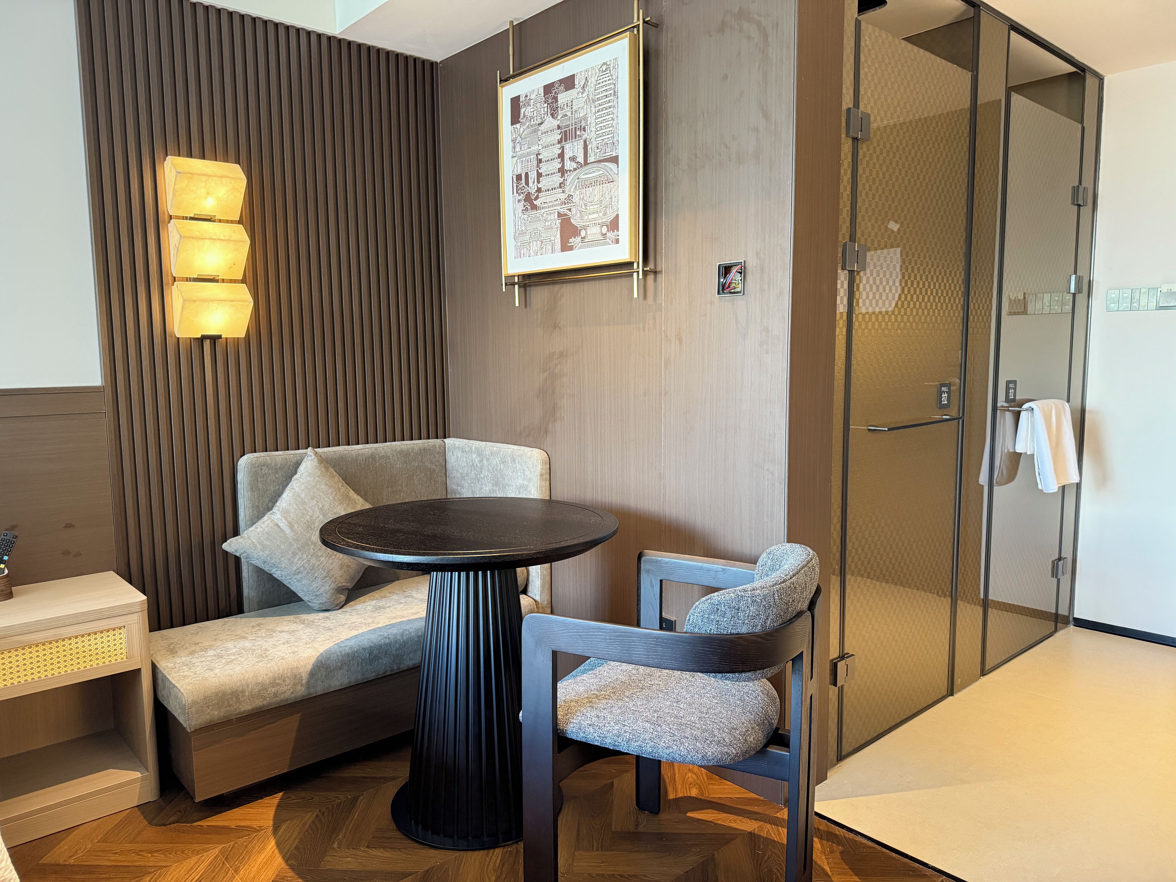 Une chambre d’hôtel moderne avec un canapé, une table ronde et une chaise. On y trouve un luminaire fixé au mur et un tableau encadré.