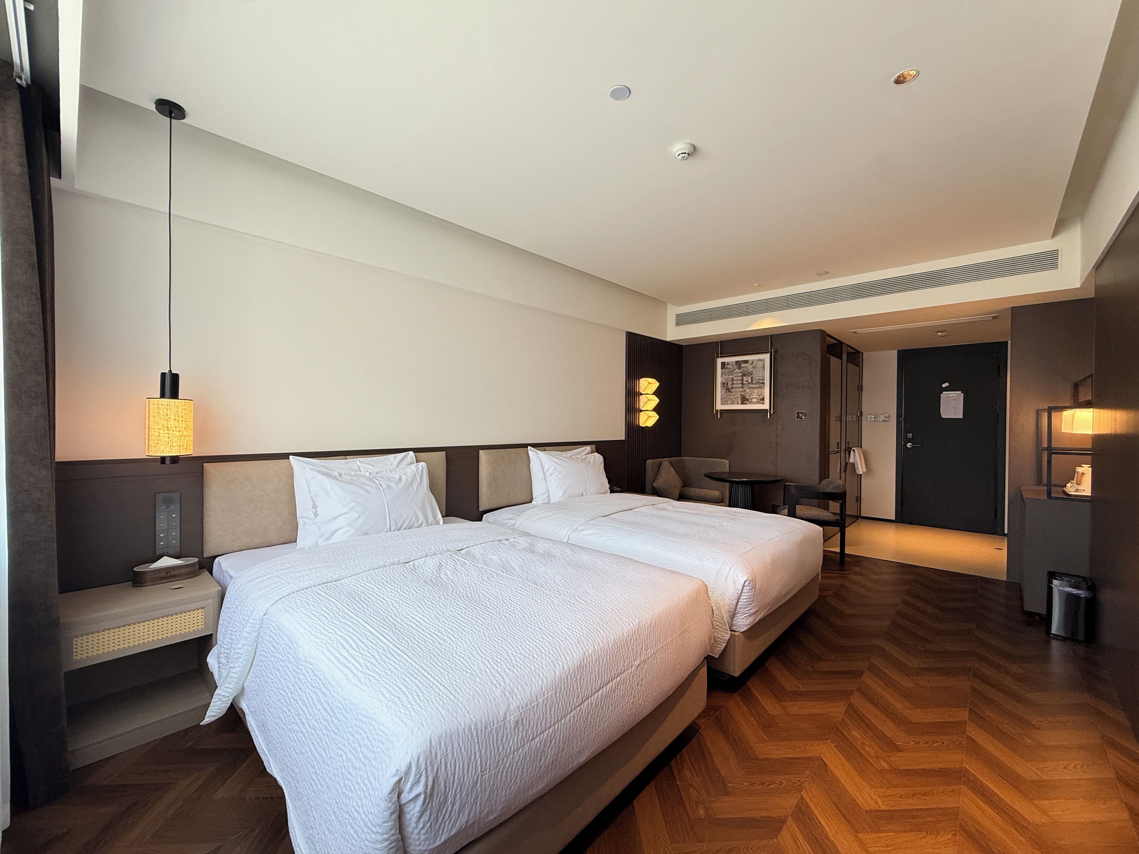 A hotel room with two beds, a desk, a chair, a lamp, and a door leading to a bathroom.