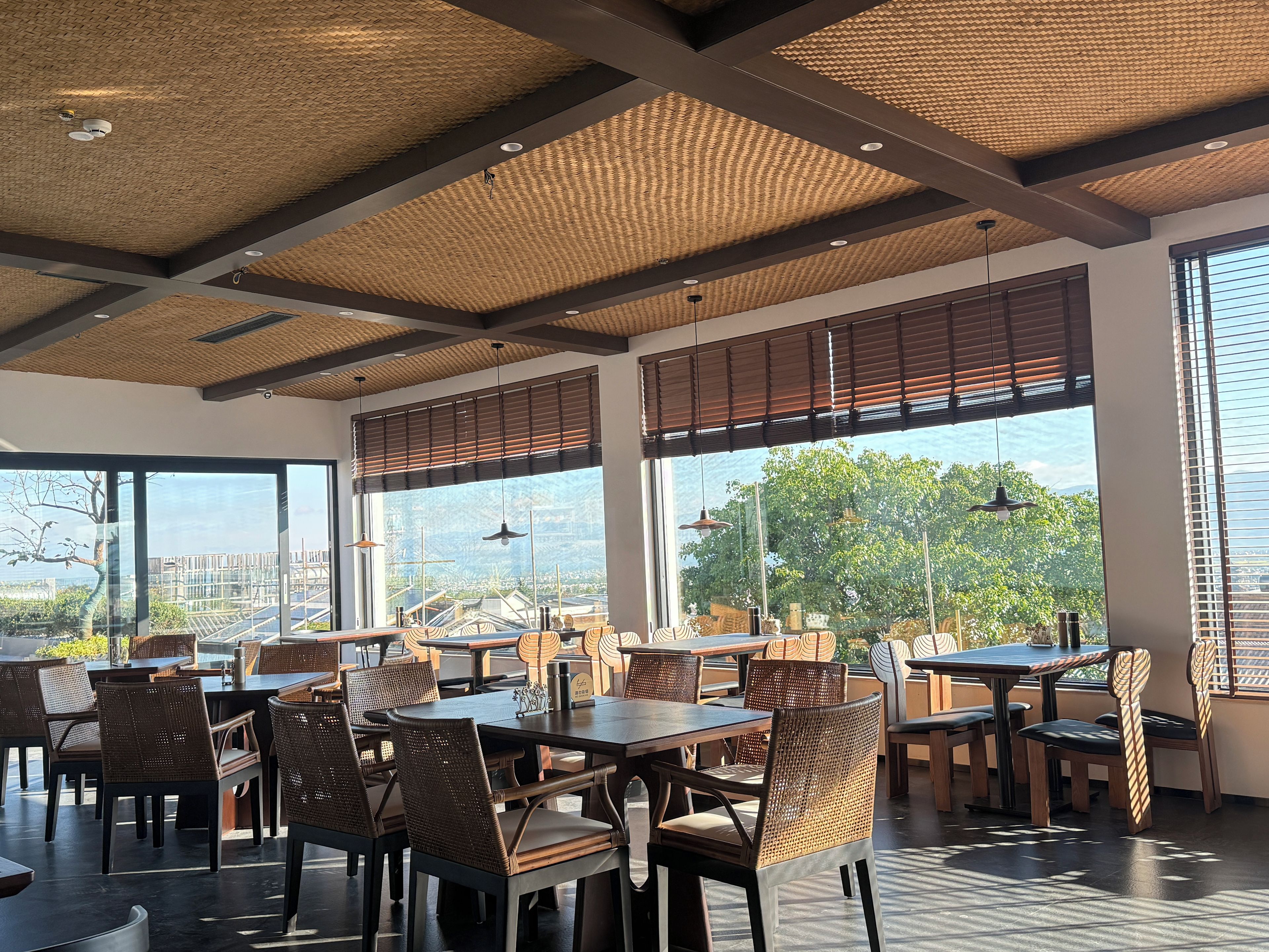 Un restaurant doté de hauts plafonds, de panneaux décoratifs en fibres tressées et de grandes fenêtres offrant une vue sur la verdure et les bâtiments. Les tables et les chaises sont disposées pour accueillir les clients.