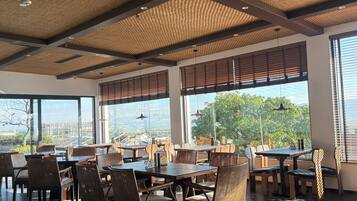Un restaurant doté de hauts plafonds, de panneaux décoratifs en fibres tressées et de grandes fenêtres offrant une vue sur la verdure et les bâtiments. Les tables et les chaises sont disposées pour accueillir les clients.