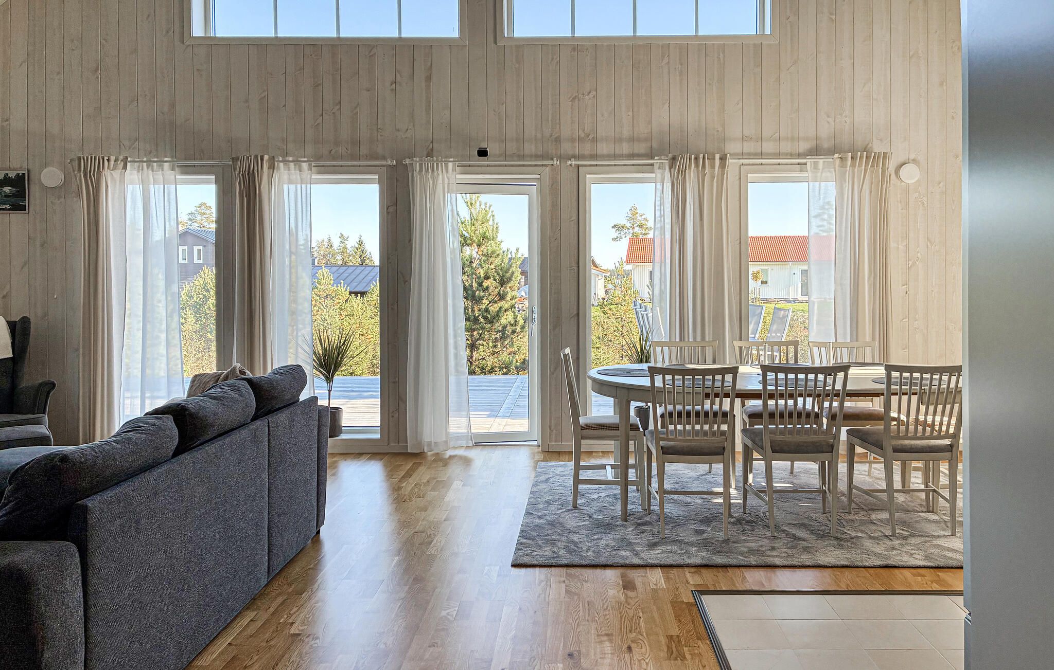 A spacious living room with a grey sofa, a dining table with chairs, and large windows with curtains.