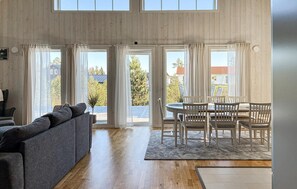 Un salón espacioso con un sofá gris, una mesa de comedor con sillas y grandes ventanas con cortinas.