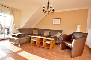 A living room with a sofa, a wooden coffee table, a brown leather armchair, a chandelier, and a large window with curtains. - Holiday apartment with 2 bedrooms (Norddeich)