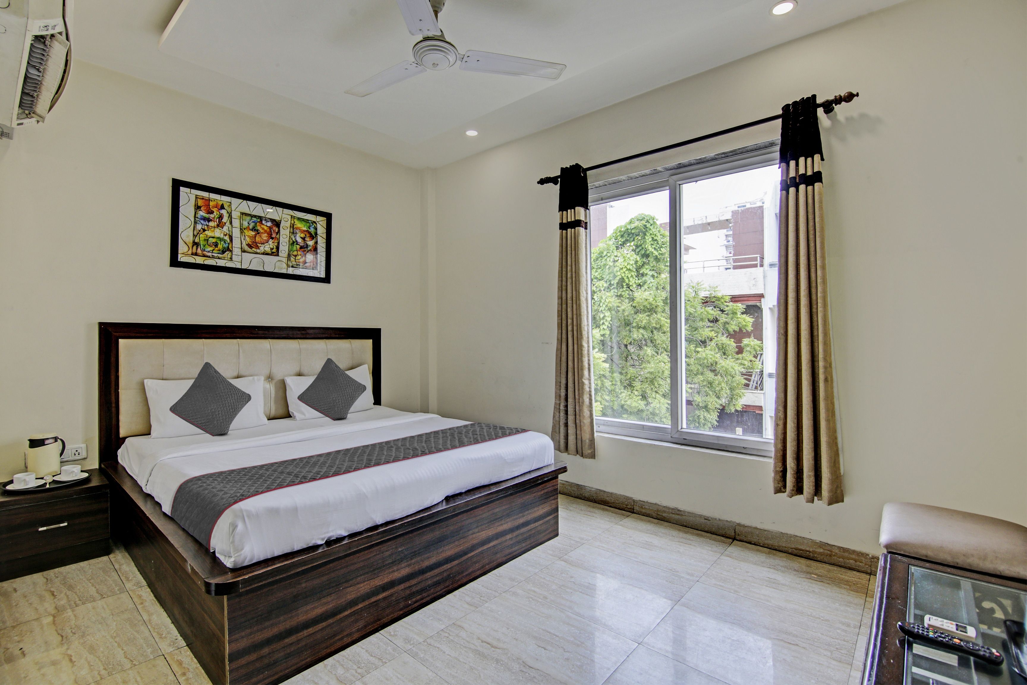 A hotel room with a bed, a nightstand, a window with curtains, a ceiling fan, and a framed picture on the wall.
