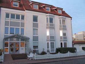 Ein mehrstöckiges Gebäude mit weißer Fassade, Hausnummer und einer kleinen Veranda.