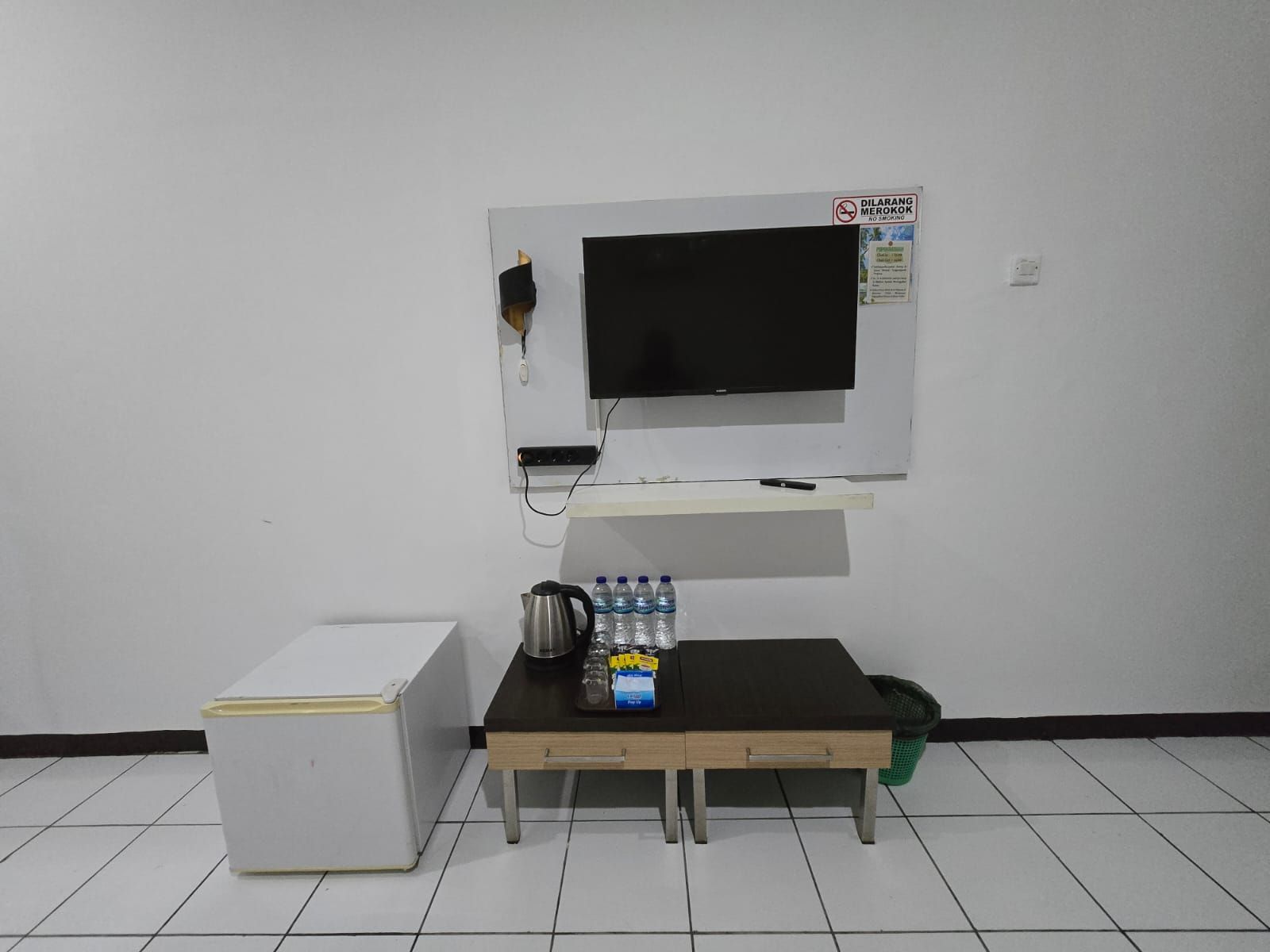 Una habitación con un refrigerador blanco, una mesita con una tetera, botellas de agua y un televisor montado en la pared.