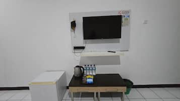 Una habitación con un refrigerador blanco, una mesita con una tetera, botellas de agua y un televisor montado en la pared.